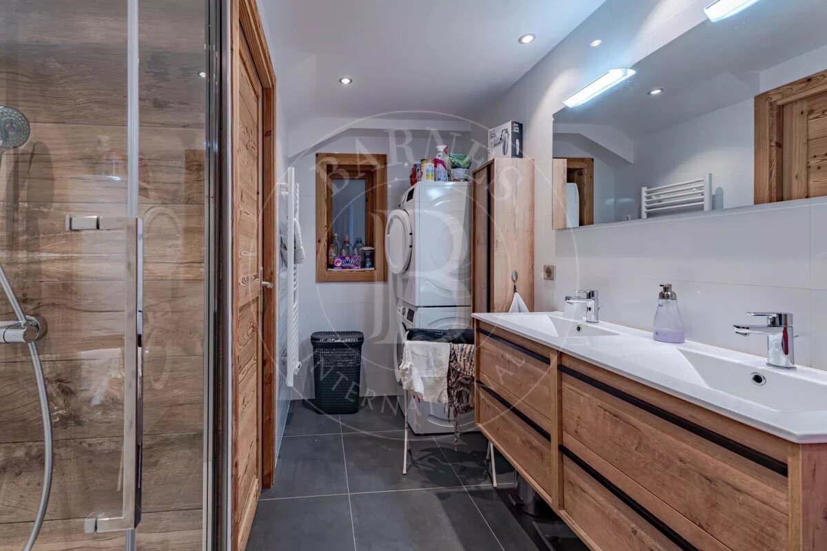Modern bathroom featuring a shower with wood-effect tiles, a double wooden vanity, and stacked washer/dryer units. Dark floor tiles complete the look.