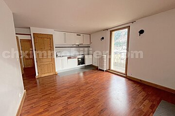 Bright studio with a white integrated kitchen, warm wooden floor, and a large glass door leading to a balcony with mountain views.