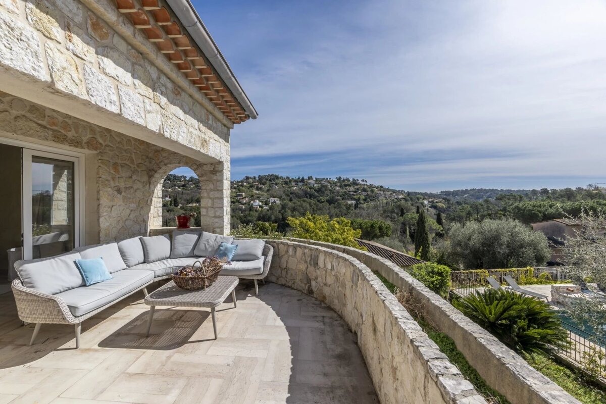A stone terrace with a curved wicker sofa overlooking a lush green valley under a bright blue sky.