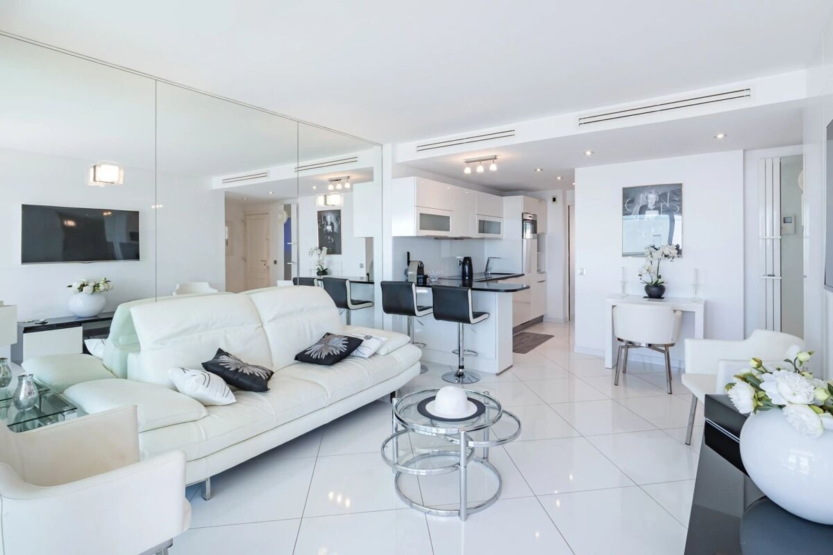 Modern white apartment interior featuring a sleek leather sofa, tiled flooring, a mirrored wall, and an open kitchen with bar stools.