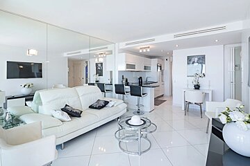 Modern white apartment interior featuring a sleek leather sofa, tiled flooring, a mirrored wall, and an open kitchen with bar stools.