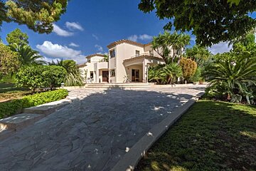 A large beige Mediterranean-style house with a stone driveway, lush green garden, and palm trees under a sunny blue sky.