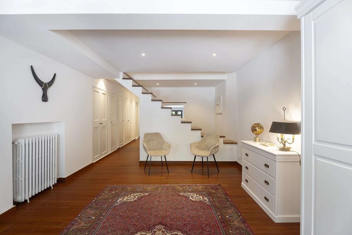 An elegant white hallway featuring wooden floors, a red rug, floating stairs, two modern chairs, built-in storage, and a dresser with a lamp.