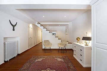 An elegant white hallway featuring wooden floors, a red rug, floating stairs, two modern chairs, built-in storage, and a dresser with a lamp.