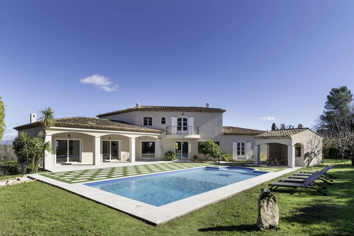 A beautiful Mediterranean-style villa with a swimming pool, green lawn, and sun loungers under a clear blue sky.