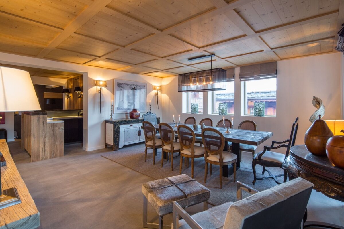 Elegant dining room with a long wooden table, chic chandelier, cozy upholstered chairs, and wood-paneled ceiling, connected to a kitchen area.