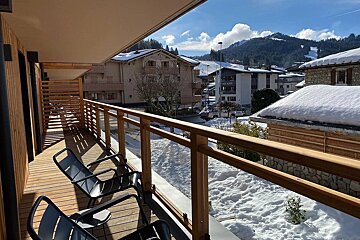 Modern wooden balcony with chairs overlooks a sunny, snow-covered village, buildings, and distant ski slopes on mountains.