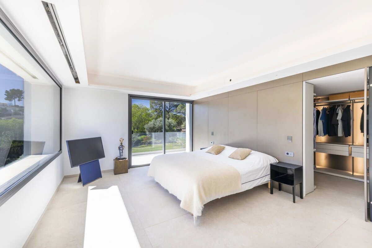 A bright, modern bedroom with large windows overlooking a lush garden. Features a white bed, sleek TV, and an open walk-in closet.