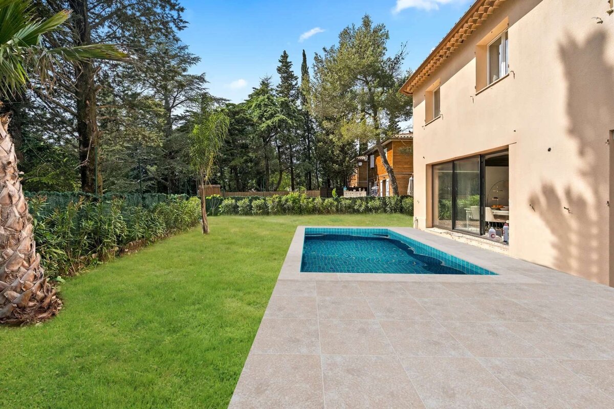 A modern house features a small blue swimming pool and a paved patio, alongside a lush green lawn bordered by dense trees under a clear blue sky.