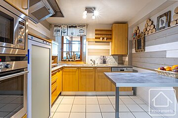A warm, light-wood kitchen with stainless steel appliances, white tiled floor, and decorative shiplap-style walls. Features include a window, sink, and small island.