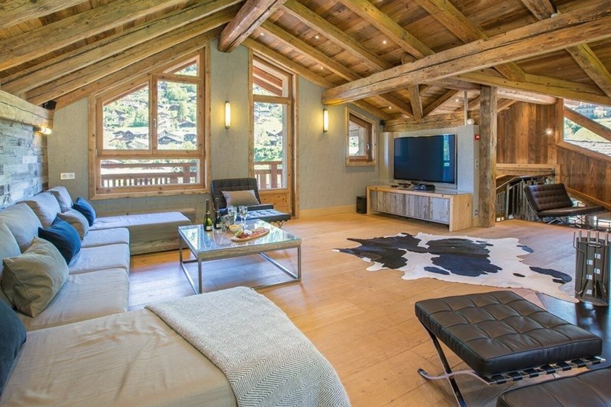 Cozy alpine chalet living room featuring exposed wooden beams, large windows with scenic views, a cowhide rug, and modern furniture.