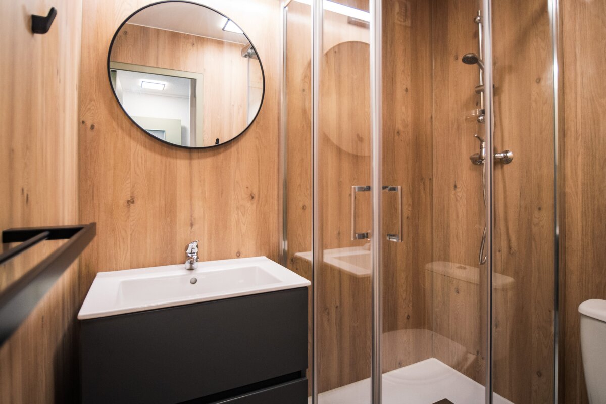A modern bathroom with warm wood-paneled walls, a white sink on a dark vanity, a round mirror, and a glass shower enclosure.