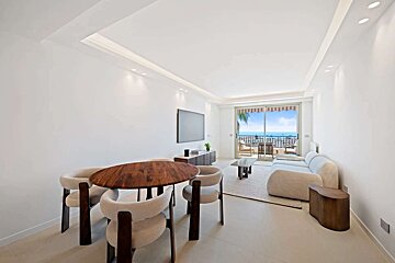 A modern, minimalist white living/dining room with a wooden table, light sofa, TV, and a balcony offering a stunning ocean view.