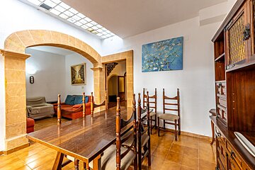 A dining room featuring a long wooden table and chairs, connected by arched doorways to a living area and a hallway with stairs. It has white walls, a tiled floor, and a skylight.