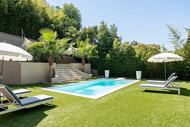 A sunny backyard featuring a swimming pool, green lawn, lounge chairs with umbrellas, palm trees, and lush green hedges.