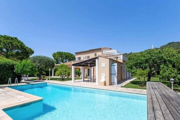 A Mediterranean-style house with a large swimming pool, surrounded by green trees and a garden, under a bright blue sky.