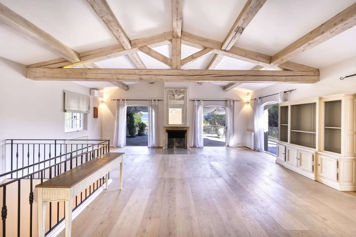 Spacious, bright room with vaulted wood-beamed ceiling, light wooden floors, fireplace, and arched glass doors leading to outdoor areas with water views.
