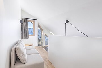 A modern, bright attic room with a light sofa, large windows showcasing snowy mountains, white walls, and a minimalist black lamp.