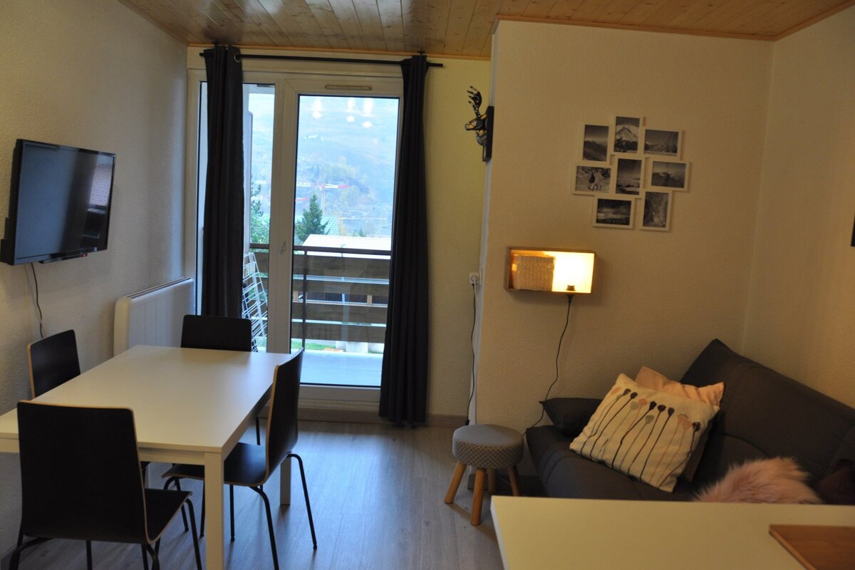 A modern living space with a dining table, wall-mounted TV, sofa, and a balcony offering an outdoor view, all under a wooden ceiling.