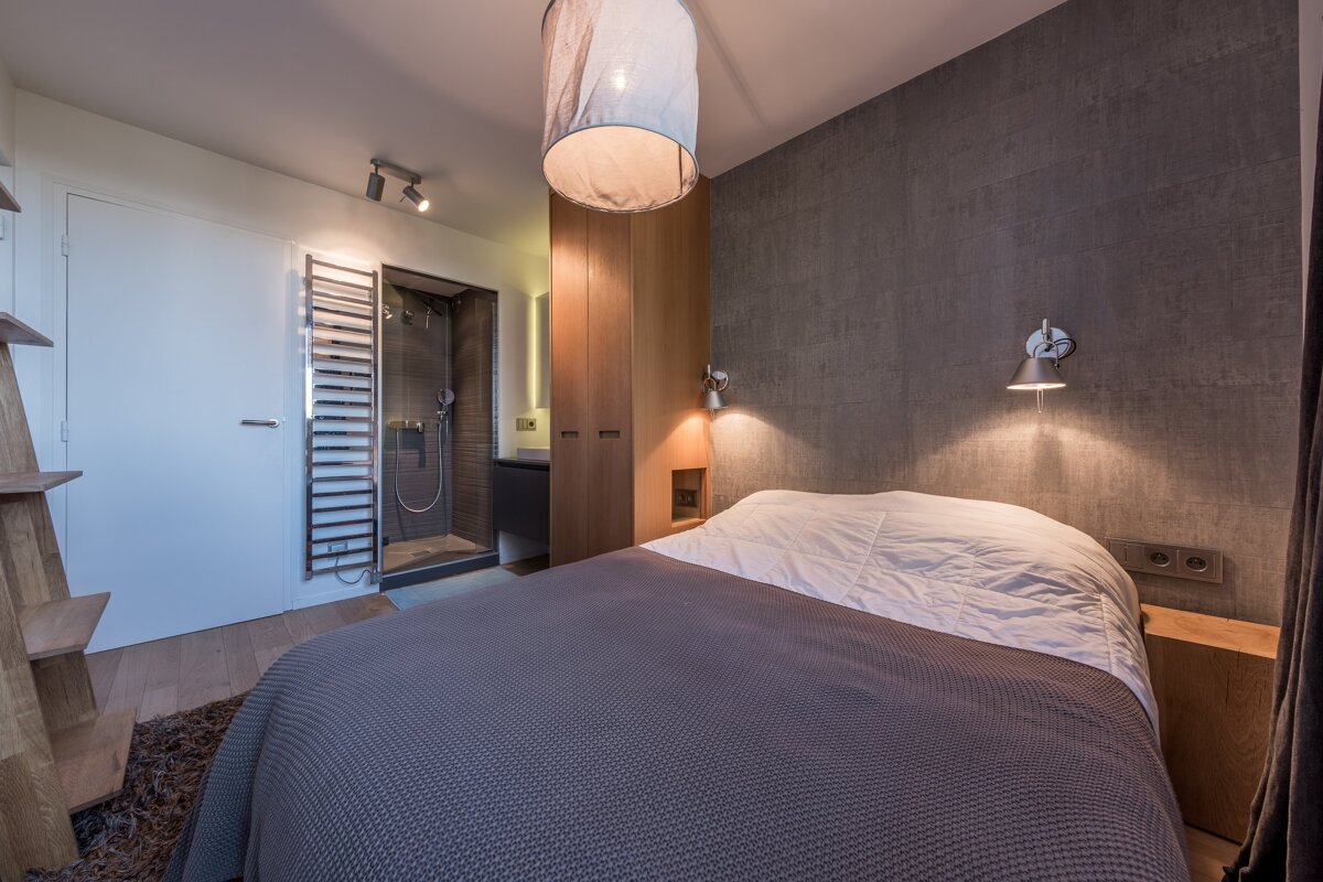 Modern bedroom features a bed, grey accent wall, and an open glass shower. Warm lighting from various fixtures creates a cozy and integrated space.
