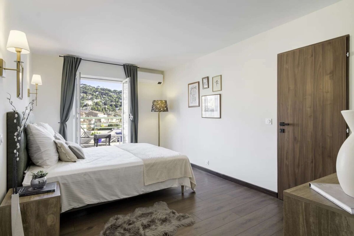 A bright, modern bedroom with a bed, wooden floor, and white walls. French doors open to a balcony with a scenic view of a hillside town.