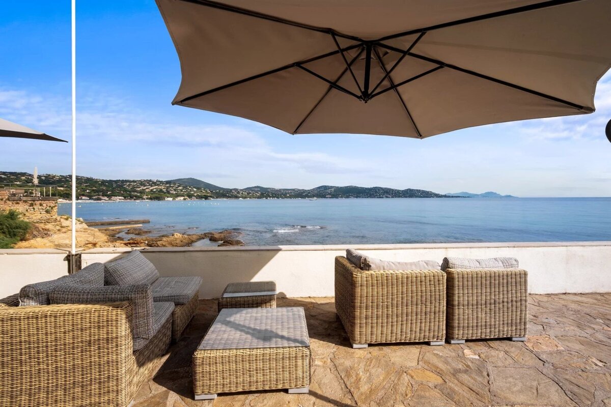 Patio with wicker furniture under a large umbrella, offering a scenic view of the blue sea and distant hills.