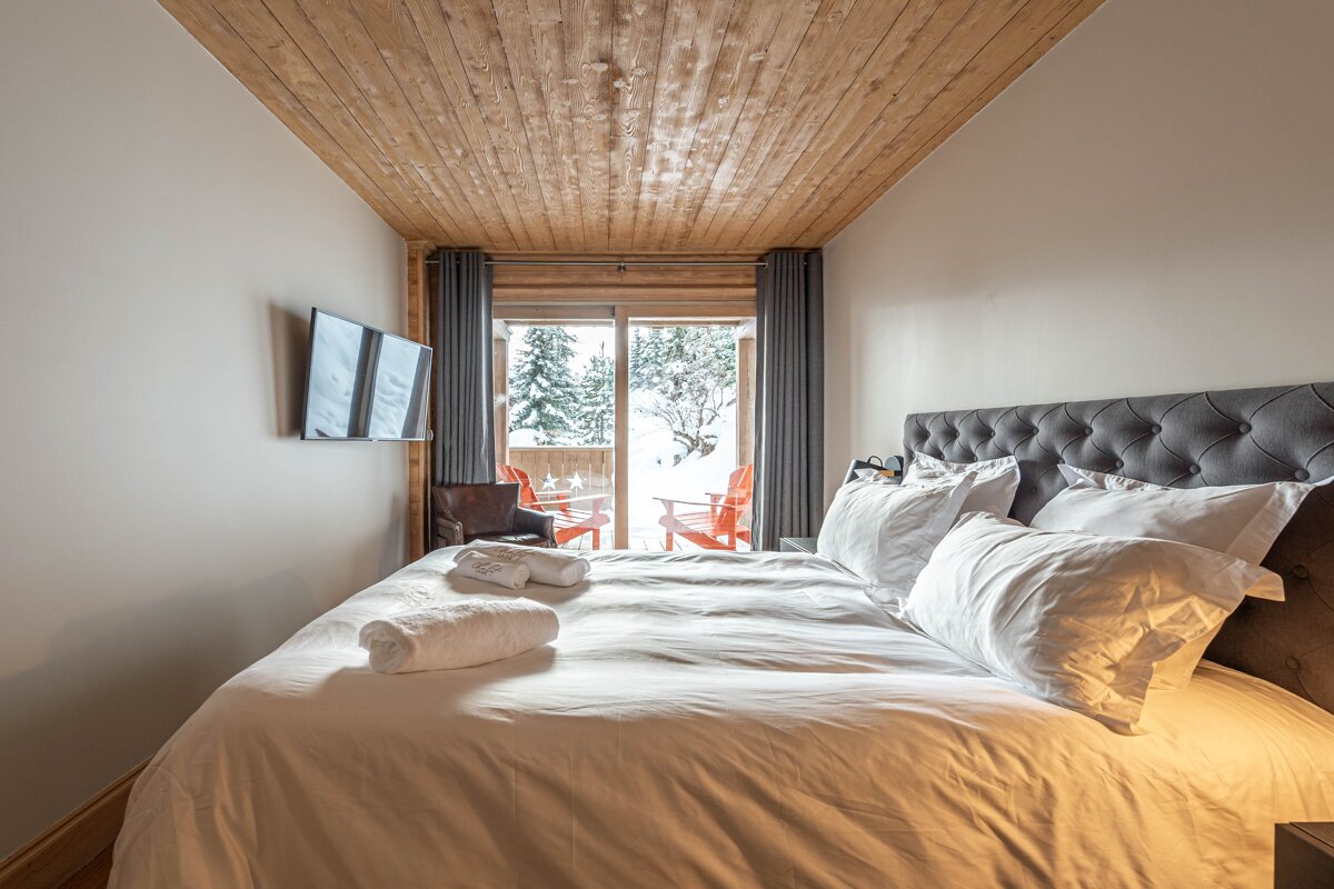 Cozy chalet bedroom with a wooden ceiling, large white-clothed bed, and glass doors looking out onto a snowy balcony.