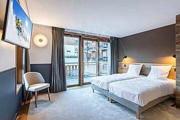 A modern bedroom with two twin beds, wooden accents, a wall-mounted TV showing skiing, an armchair, and large glass doors leading to a balcony.