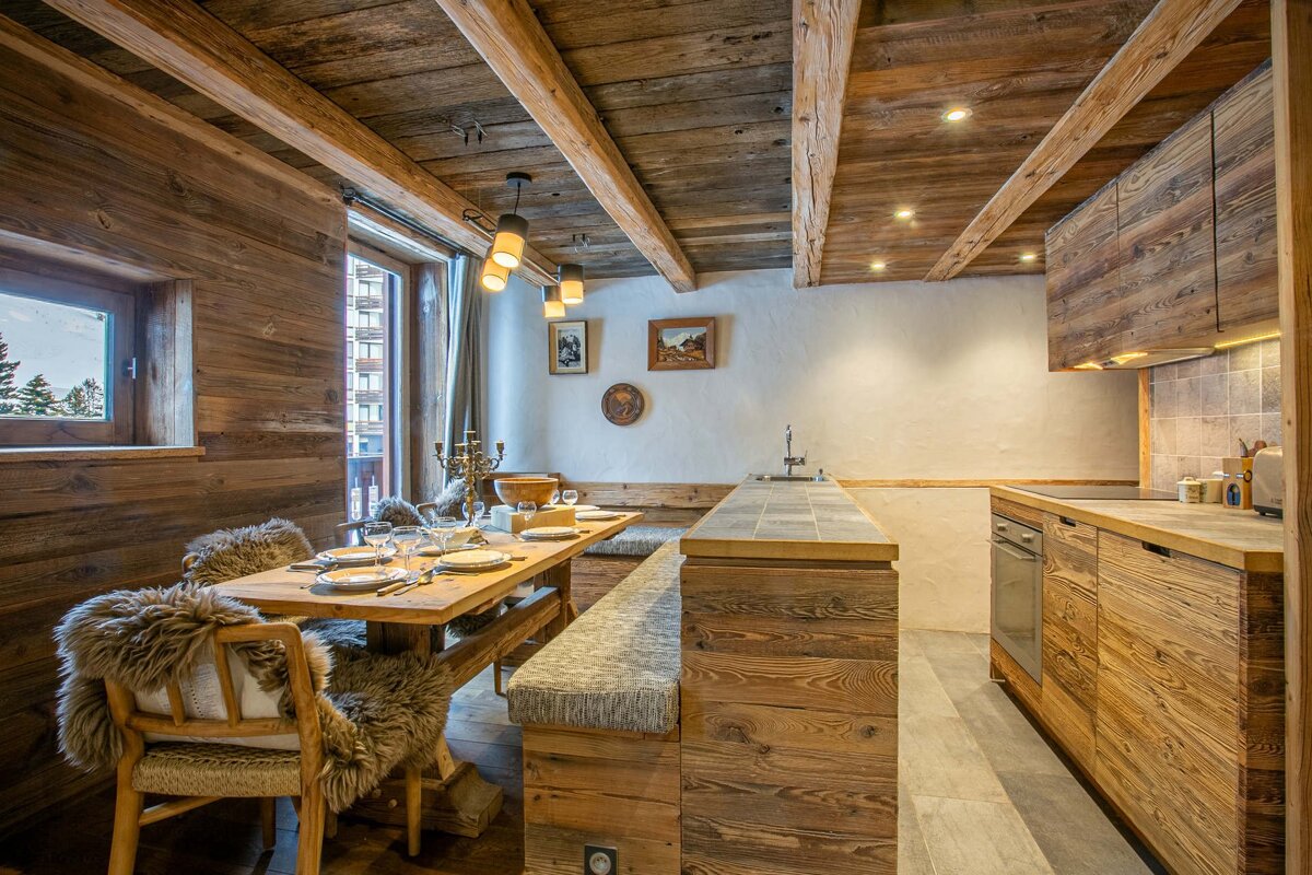 A rustic, cozy chalet interior featuring a wooden dining area with furry chairs, a set table, and a kitchen with wooden cabinets and modern appliances.
