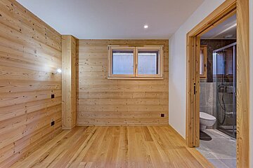 Empty room with light wood paneling and floors, featuring a window and an open doorway leading to a modern bathroom.