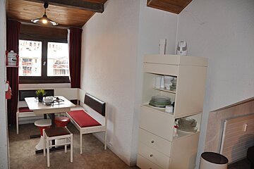 A cozy dining nook features a white table, red-cushioned benches, and stools by a window with red curtains. A dish cabinet is nearby, all under a wooden ceiling.