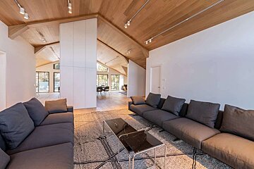 Modern living room with a vaulted wood ceiling, white walls, dark gray sofas, and a geometric rug. An open layout reveals a dining area.