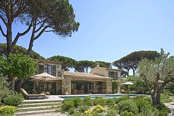 A luxury stone villa with a swimming pool, surrounded by Mediterranean pines and olive trees under a clear blue sky.