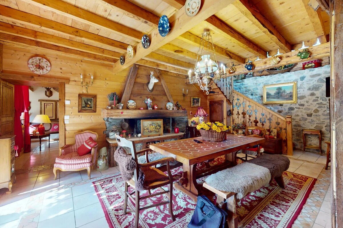 Cozy, rustic cabin interior featuring a wooden dining table, stone fireplace, decorative ceiling beams, and a patterned rug.