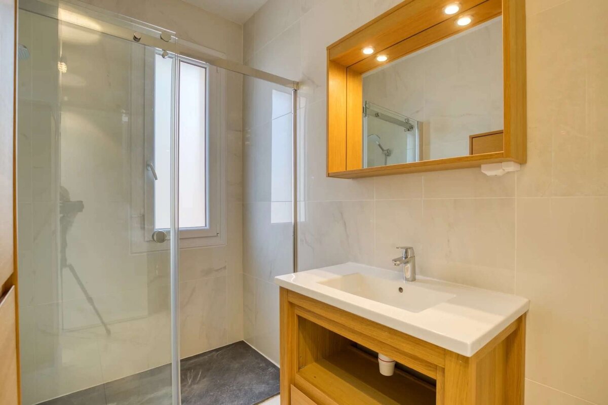 Modern bathroom with a glass shower enclosure, wooden vanity and lighted mirror, and light tiled walls.