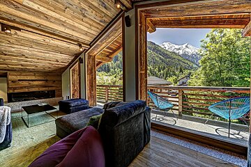 A modern, wood-clad chalet living room featuring a balcony with blue chairs and panoramic views of snow-capped mountains and lush green forests.