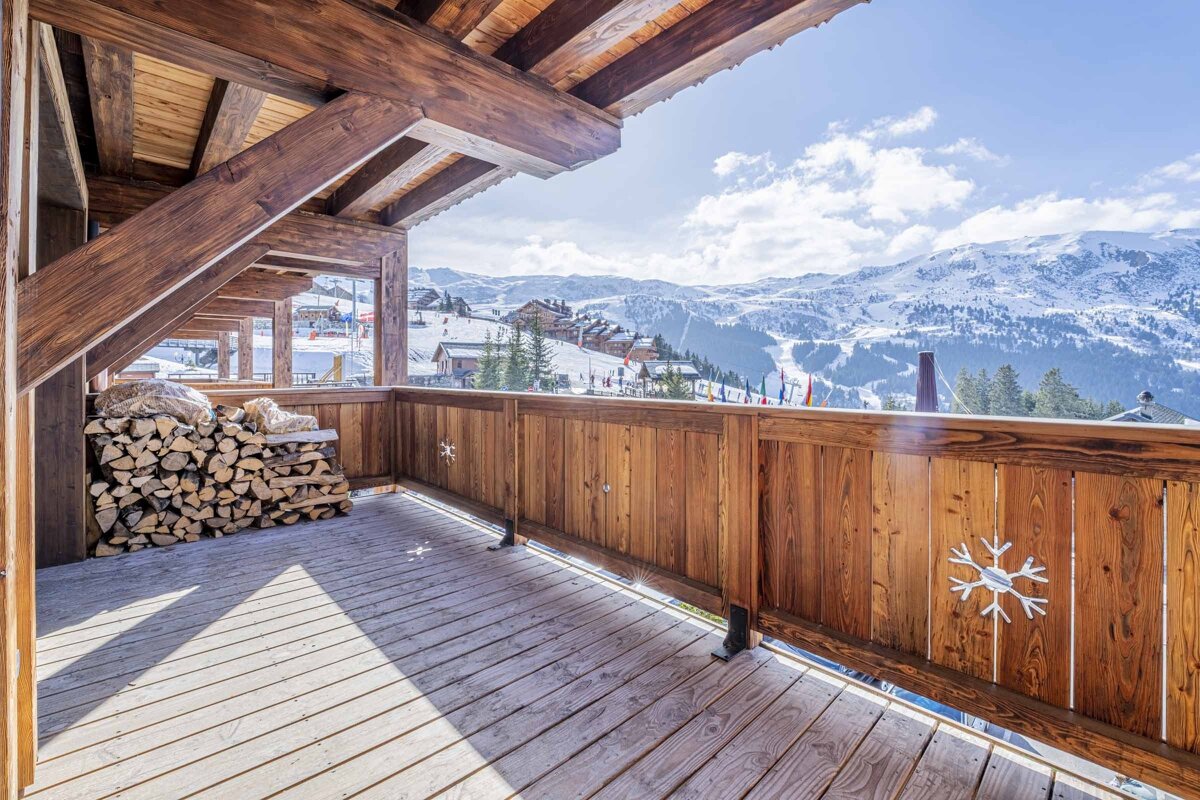 A cozy wooden balcony with firewood overlooks a sunny snowy mountain ski resort, decorated with a snowflake on the railing.