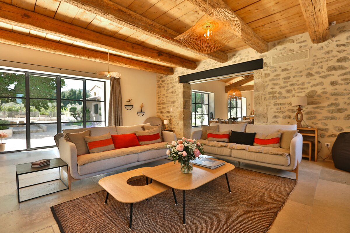 A rustic yet modern living room features exposed stone walls, wooden beamed ceilings, large windows, light sofas with colorful cushions, and woven pendant lights.