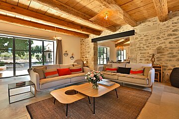 A rustic yet modern living room features exposed stone walls, wooden beamed ceilings, large windows, light sofas with colorful cushions, and woven pendant lights.