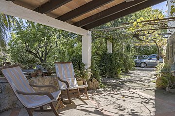 A sunny outdoor patio with two striped rocking chairs under a leafy pergola. Lush greenery surrounds the area, with cars visible in the distance.