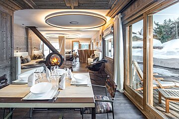 Warm, rustic modern chalet: dining table, circular fireplace, and snowy winter views through large windows.