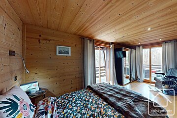 Cozy wooden bedroom with a floral bed, large windows leading to a balcony, mirrored wardrobe, and a rustic yet modern feel.