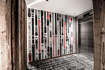A modern hallway features a wall covered in decorative skis (red, black, white), flanked by wooden panels, a metal elevator, and a patterned carpet.