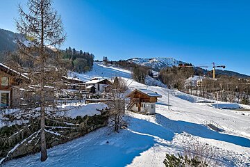 A bright, sunny winter scene of a snow-covered mountain village with chalets, ski slopes, bare trees, and distant peaks under a clear blue sky.