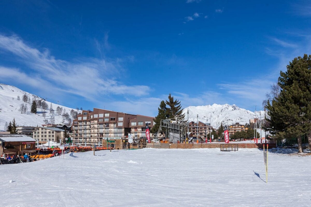 A sunny ski resort features snow-covered slopes, modern buildings, and distant snow-capped mountains against a clear blue sky.