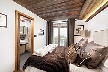 A cozy, rustic bedroom with a wooden ceiling, a large bed with brown bedding, and an en-suite bathroom visible through an open door. A window shows a snowy outdoor scene.