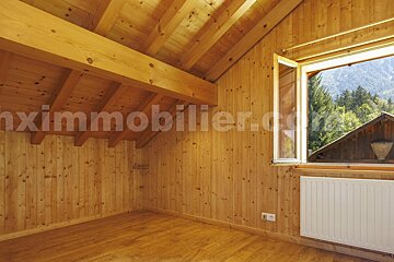 An inviting, wood-paneled room with a sloped, beamed ceiling and matching floor. A large window reveals a stunning mountain and forest landscape.