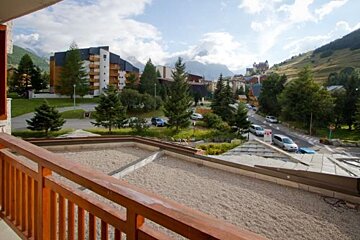 View from a wooden balcony overlooking a mountain resort village with pine trees and cloudy hills.