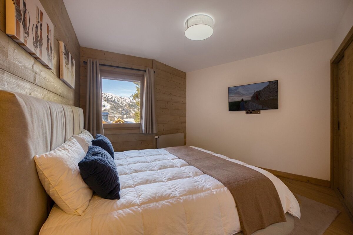 Cozy chalet bedroom with a large bed, wood-paneled walls, a wall-mounted TV, and a window offering a view of snowy mountains.