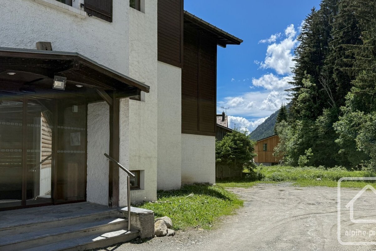 Exterior view of a chalet's entrance next to a dirt path, surrounded by trees under a blue, partly cloudy sky.